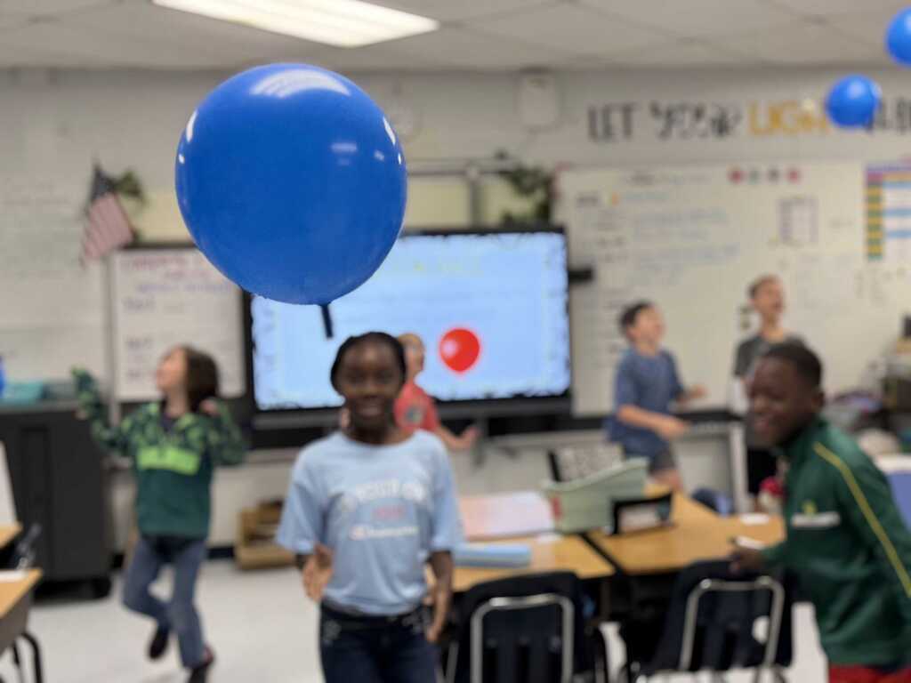 Kids in a classroom, one holding a blue balloon, with tables, chairs, and a flat screen TV behind them.