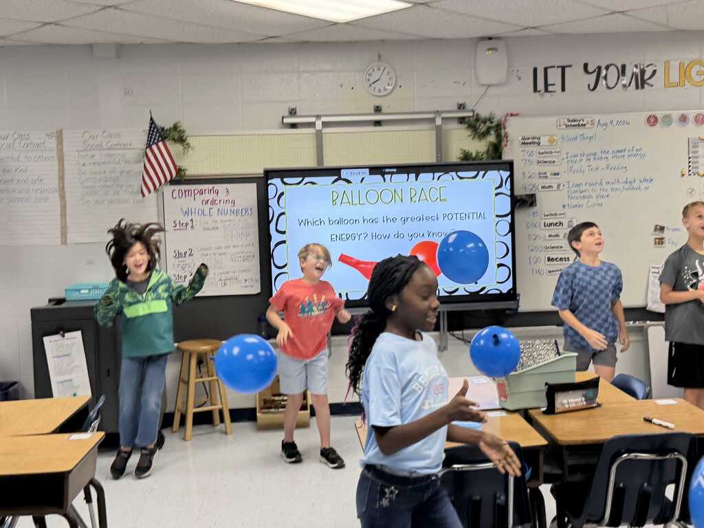 Students in a classroom, with some holding balloons, while others watch, and a large screen displays the text "BALLOON RACE"