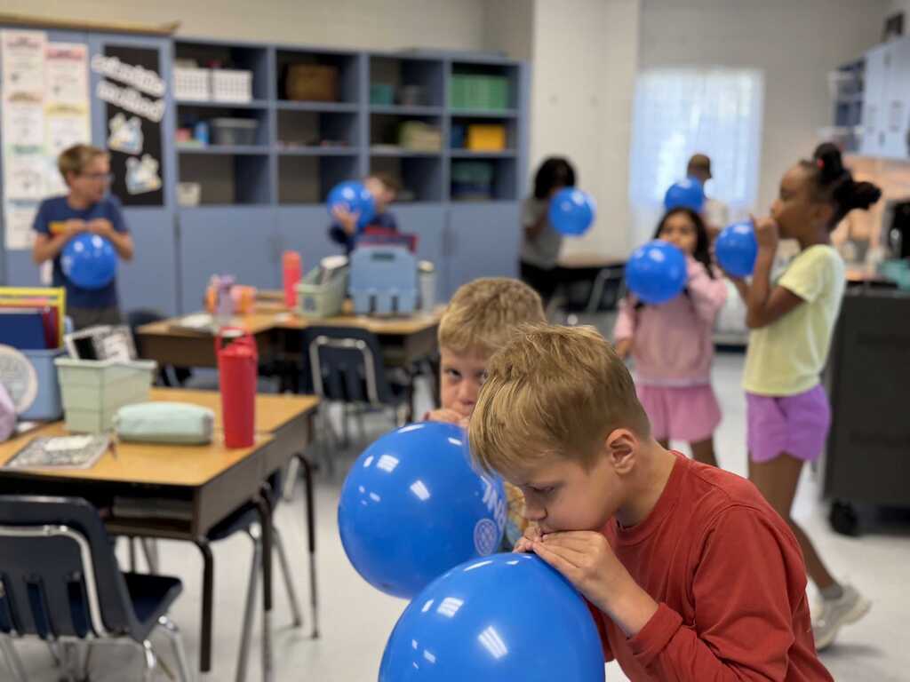 Children play with blue balloons in a classroom with desks, shelves, and a window in the background.