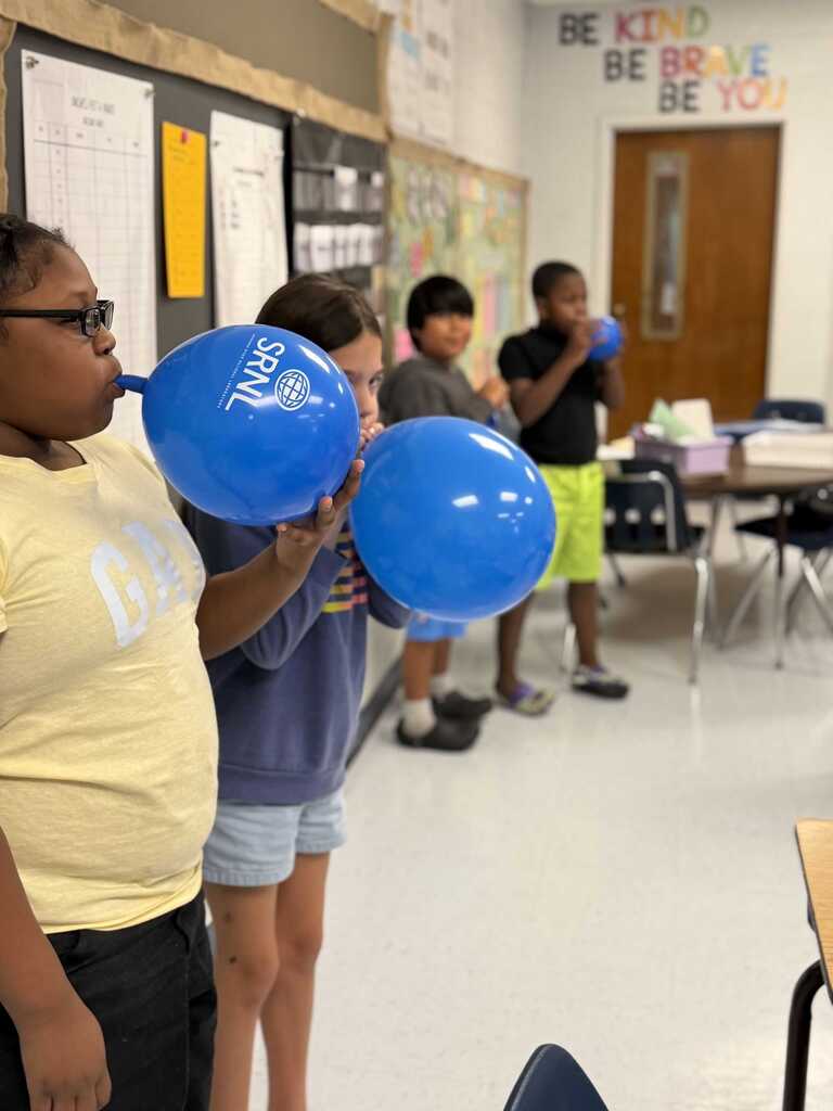 Four children stand in a classroom. Two blow up blue balloons. One holds a balloon at the mouth. Bulletin boards and a door are in the background.