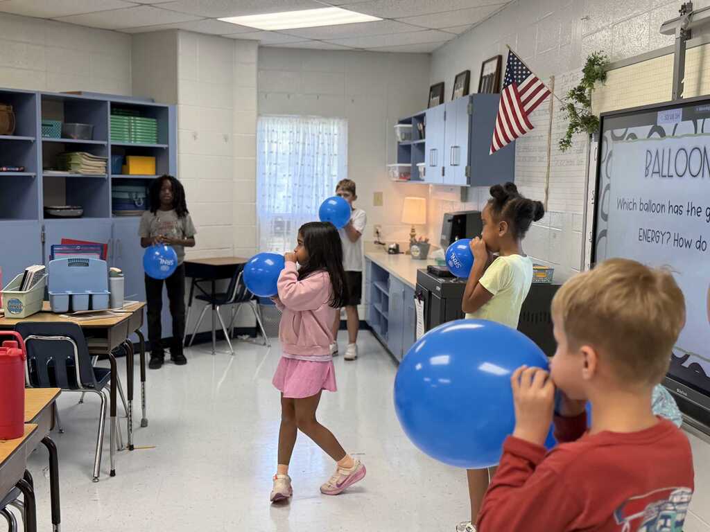 Classroom with children inflating balloons. A woman stands by the shelves, and a flag hangs on the wall.