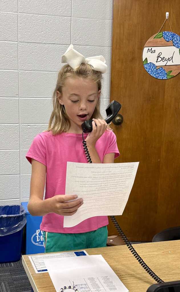 A young girl with a white bow in her hair holds a paper and phone receiver. Behind her, a door with a sign reading "Ms. Boyd" is visible.
