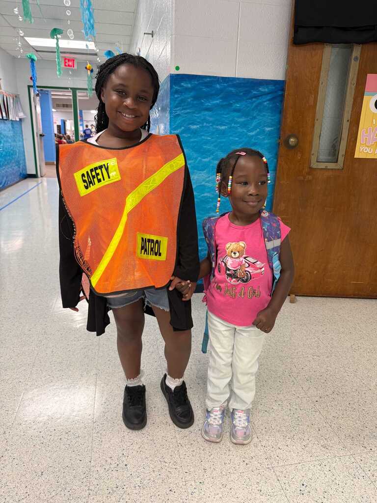 Two girls, one wearing a reflective vest and the other in a pink shirt, stand in a hallway.