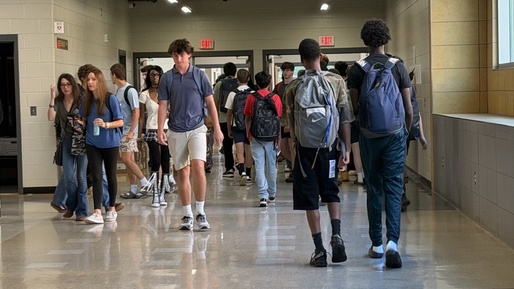 A group of students walk in a hallway with white tile flooring. Some carry backpacks.