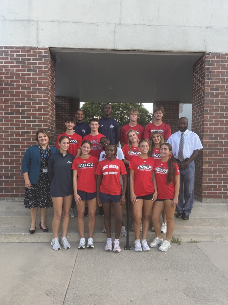 A group of athletes wearing red shirts and shorts, along with two adults, pose in front of a brick building.