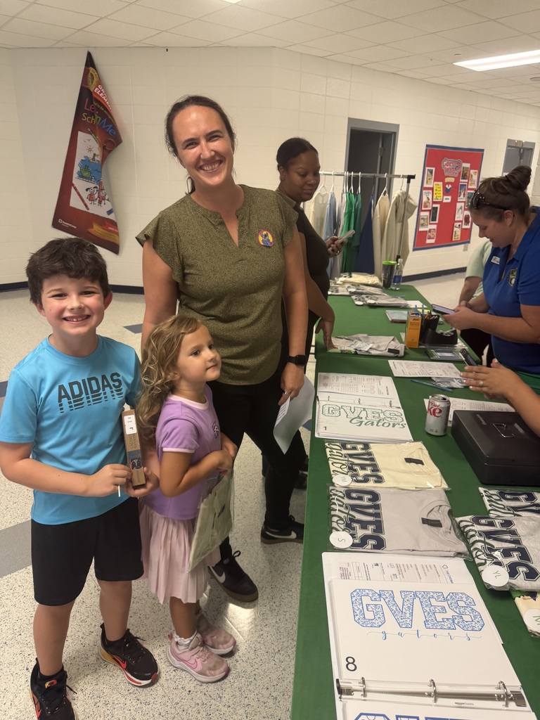 A woman and two children stand near a table covered in green cloth, with papers and a flag in the background.