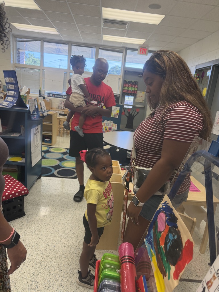 Two adults and two young children in a room with a table, rug, and shelves. One child holds a baby.