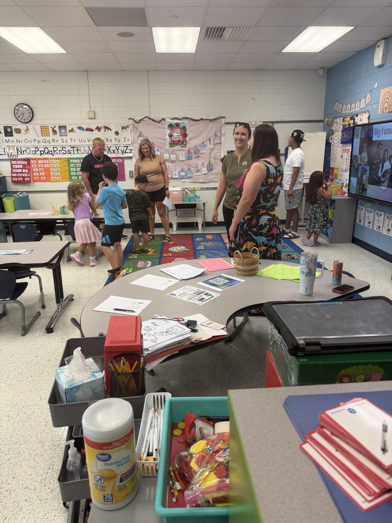 A classroom with children and adults standing around a table with papers. The room has desks, chairs, and posters on the wall.