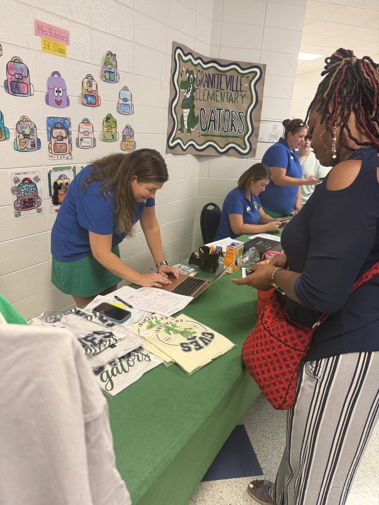 People in blue shirts work at a table covered with green fabric, surrounded by bags and papers.
