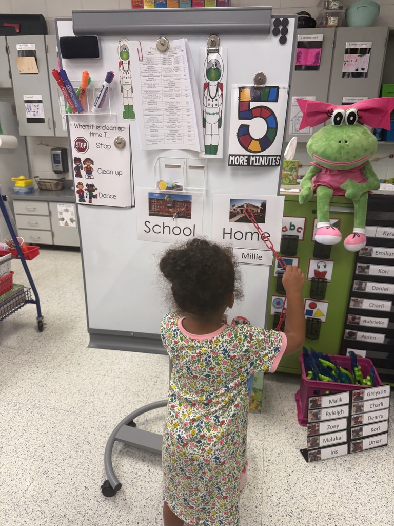A child touches a white board with numbers, letters, and a pink frog. The room has cabinets, a rolling cart, and various items.