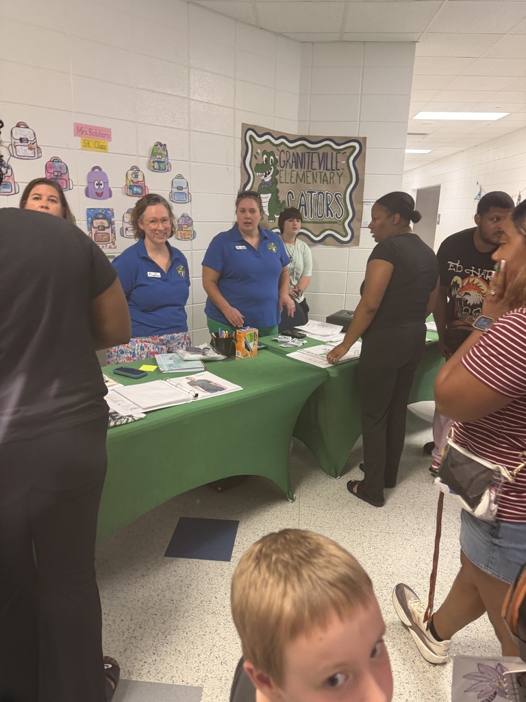 A group of people stands around tables with green covers, likely at an event. Some wear blue shirts. A boy with blonde hair looks on.