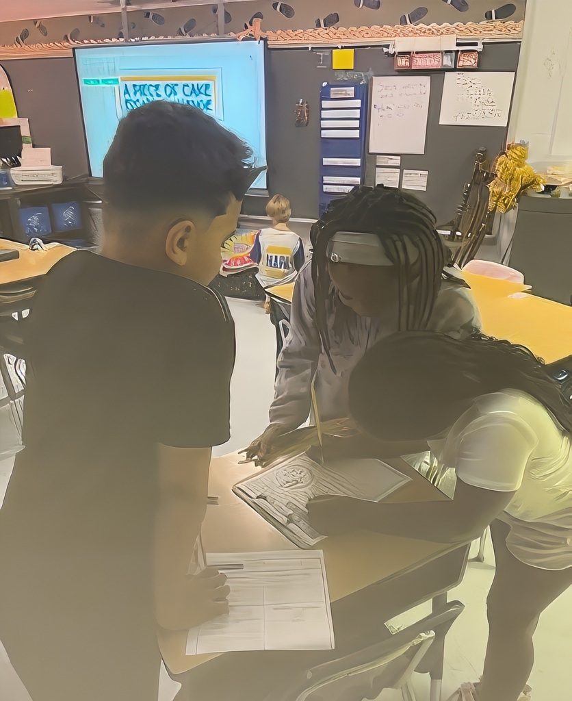 Three students in a classroom are looking at papers on a desk. A large screen shows a presentation.