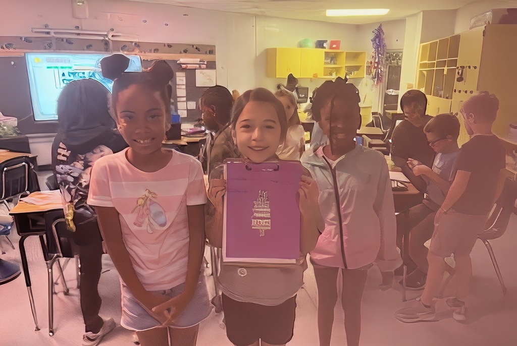Three young girls in a classroom, with one holding a purple folder, surrounded by desks and other students.