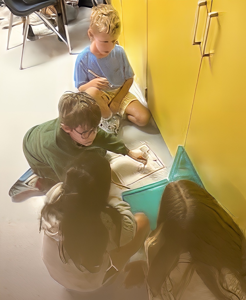 Four children in a classroom, two boys and two girls. One boy holds a paper, another works on a project.