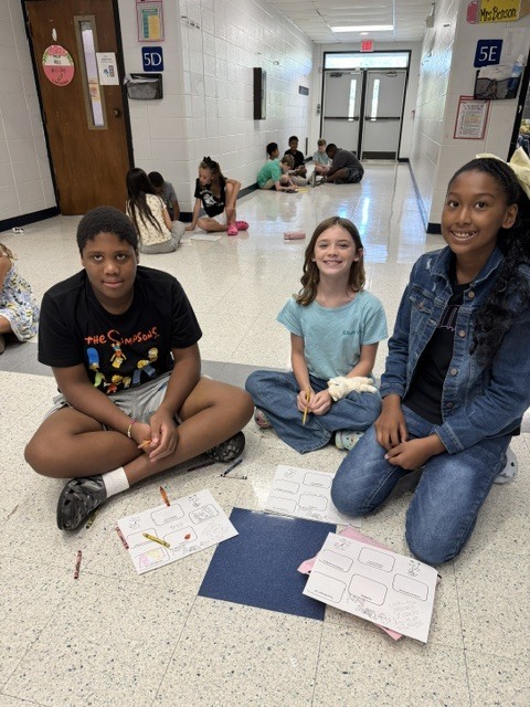 Three children, two girls and one boy, sit on the floor in a school hallway, working on papers.