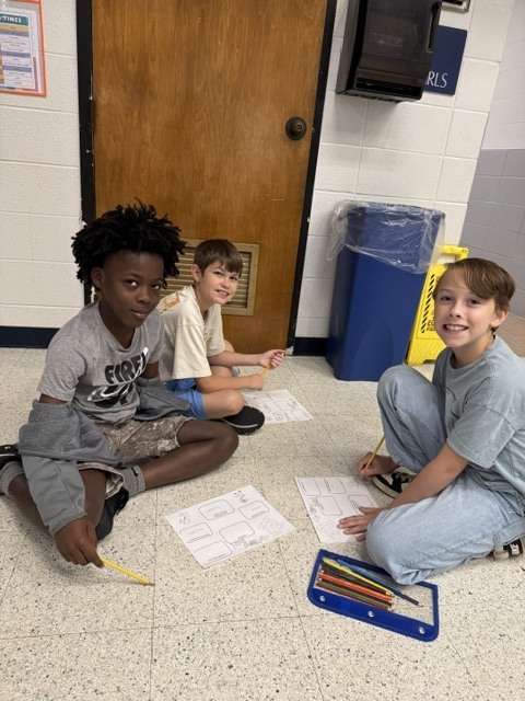 Three children sit on the floor, engaged with paper and pencils. They are in front of a door and a trash bin.