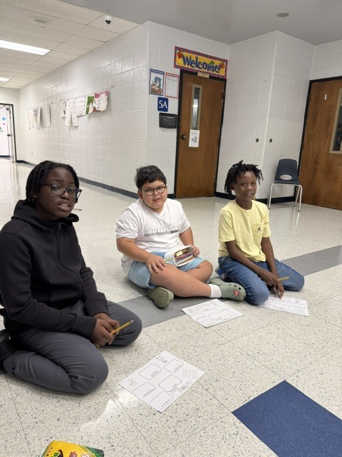Three students sitting on a floor with papers, one holding a pencil, in a hallway with white walls and doors.