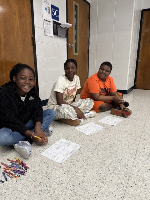 Three individuals are seated on the floor, engaging in an activity. Papers are spread out in front of them, and colored pencils are on the floor.