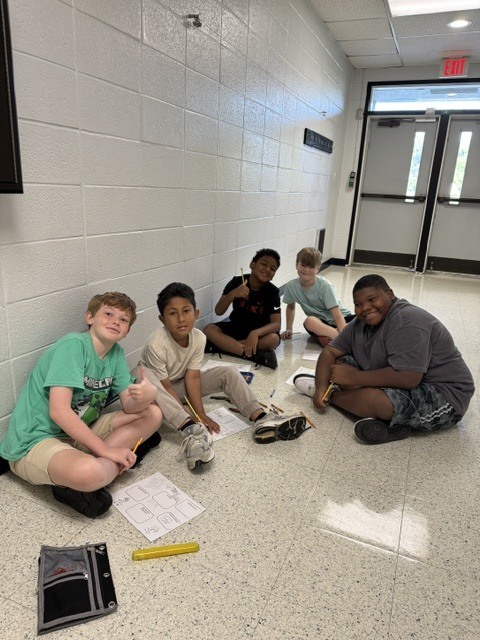 Six boys sitting on a tiled floor, engaged in a task. One holds a notebook, another has a pen.