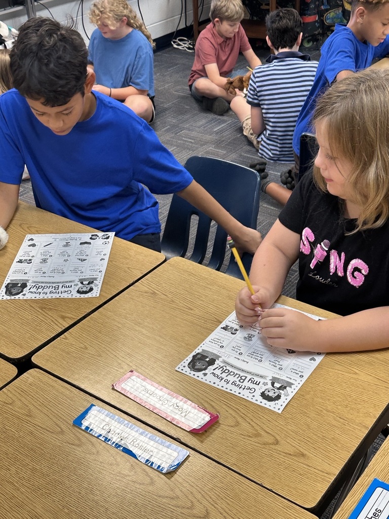 Children in a classroom sit at tables; two are working on worksheets, one holds a pencil, while others sit on the floor.