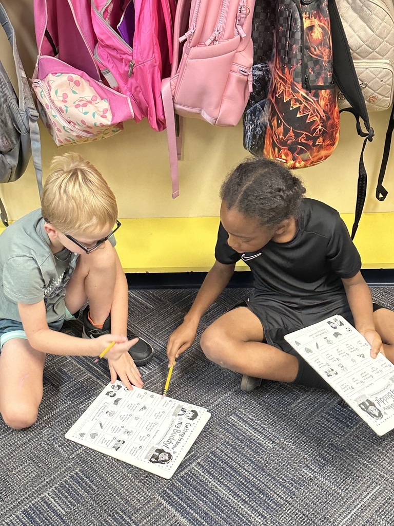 Two children sit on a floor, holding papers and writing with a pencil. One wears glasses, the other a bracelet. Various backpacks hang behind them.