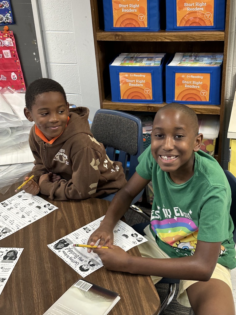 Two boys sitting at a table, smiling, working on worksheets. Behind them, a bookshelf with colorful storage boxes.