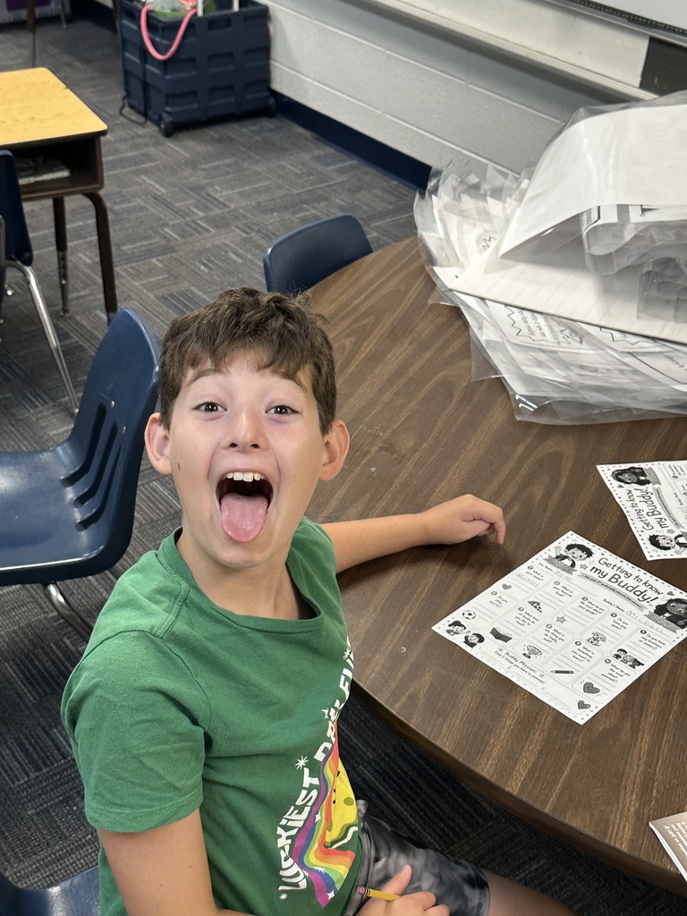 A boy in a green shirt sits at a desk with his mouth open. Behind him, desks, chairs, and papers are visible.