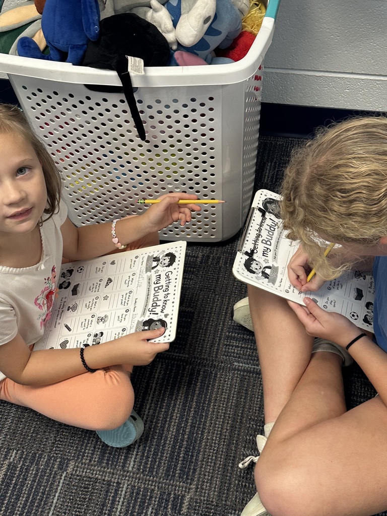 Two girls sit on a floor, writing in papers. One holds a pencil. A white laundry basket with stuffed toys is behind them.
