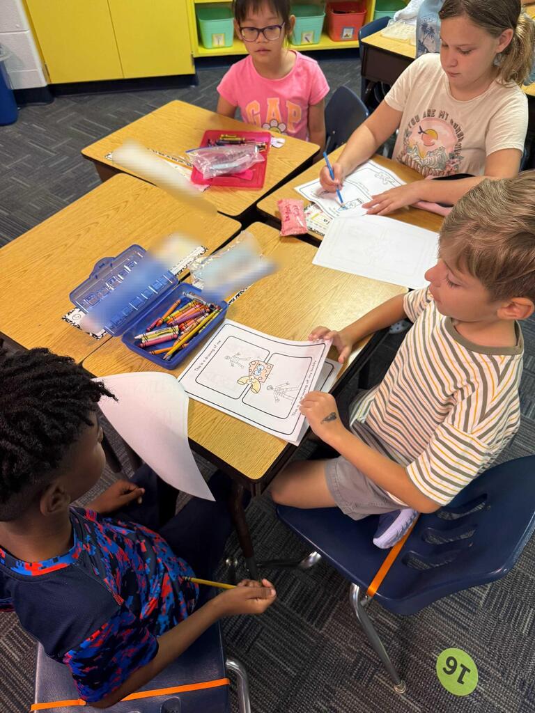Four children sit at desks in a classroom, engaged in coloring activities. One holds a paper and a pencil.