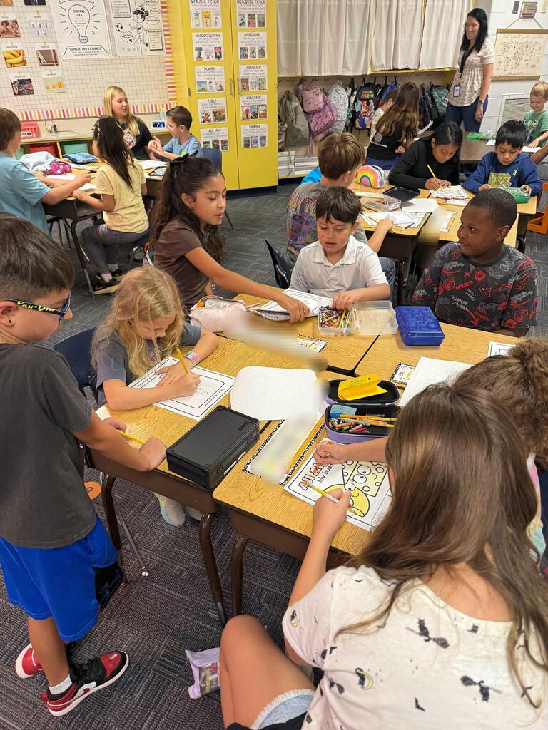 Elementary students seated at desks, engaging in various activities. A teacher is standing and observing the students.