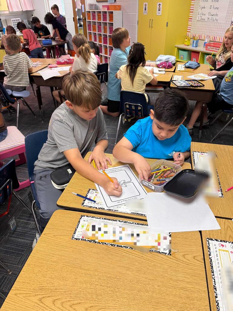Children in a classroom, engaged in an activity. Two boys sit at desks with papers and colored pencils.