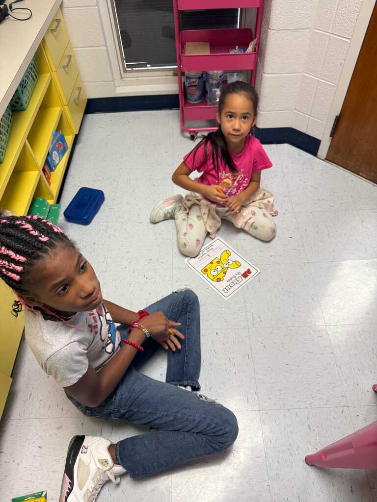 Two girls sitting on a floor, one holding a card with the letter M, a pink shelf with items, and a window behind them.