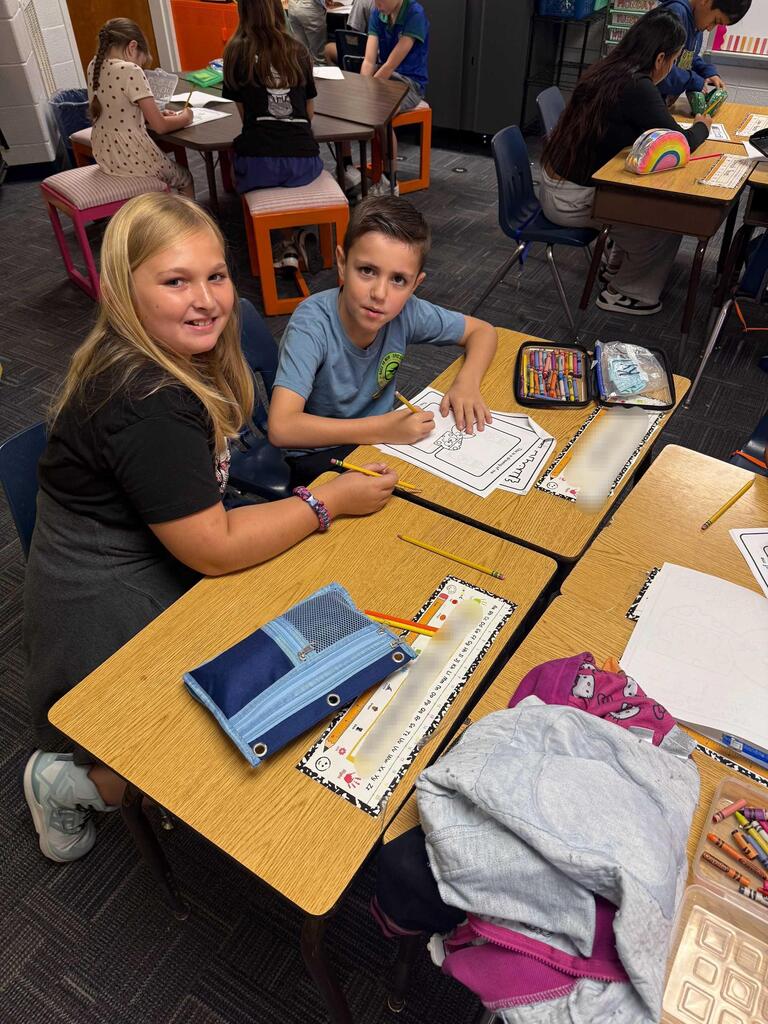 Two students in a classroom. One wears a black shirt, and the other wears a blue shirt. They sit at desks with papers, pencils, and crayons.