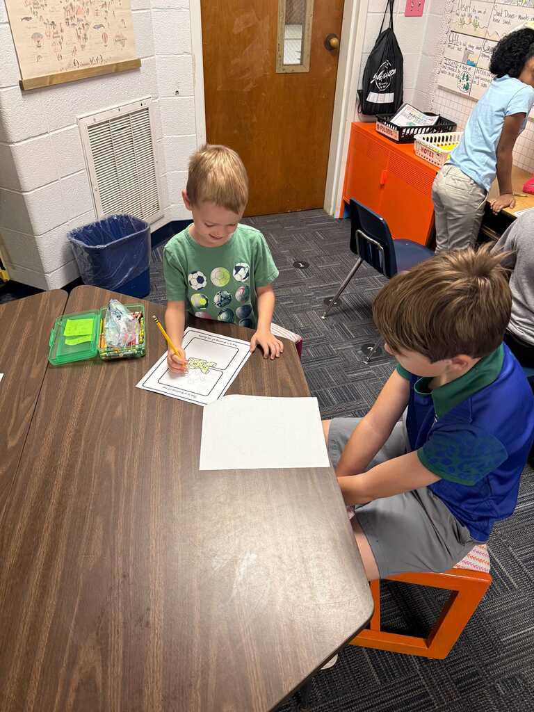 Two children seated at a desk, working on a piece of paper with pencils. Another child stands nearby.
