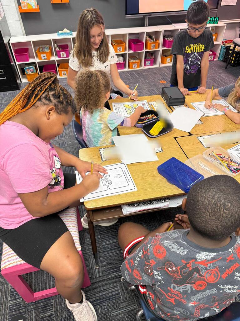 Students are seated around a table, coloring and drawing on paper. A woman stands, assisting a child.