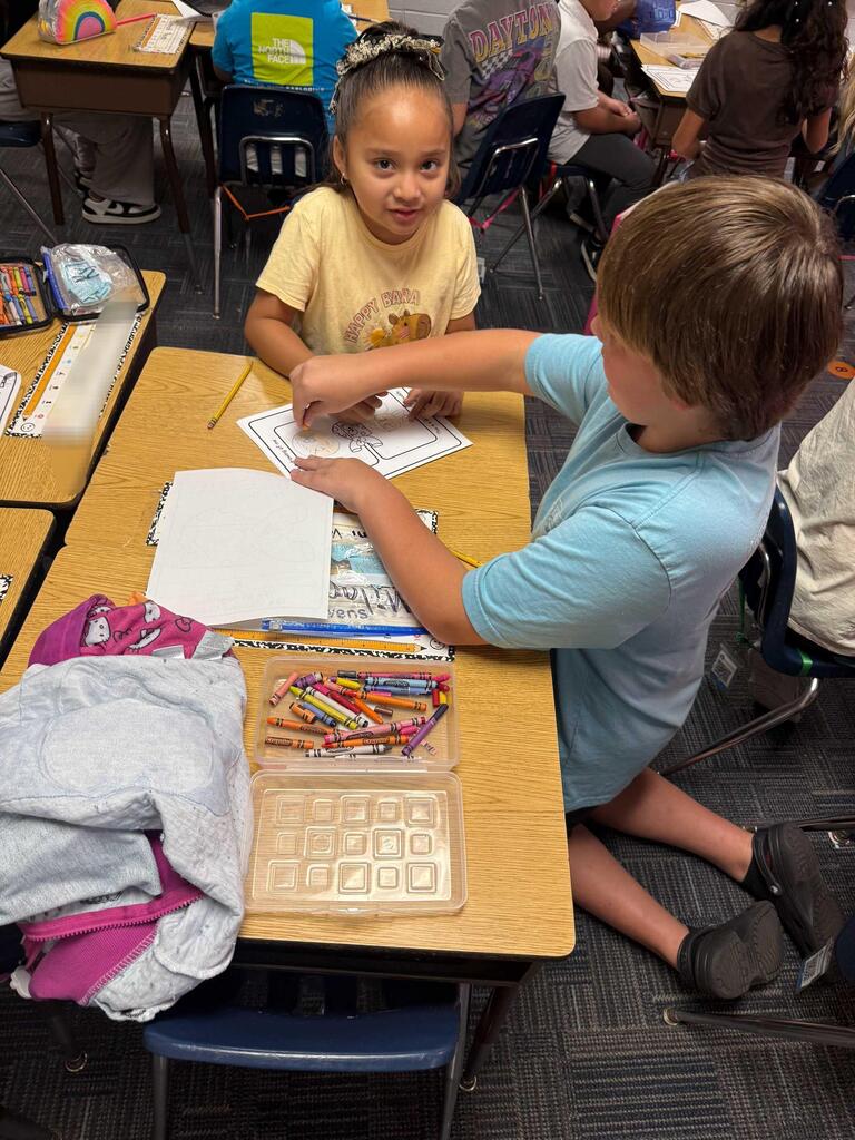 Children in a classroom setting, one using crayons, the other holding a piece of paper.