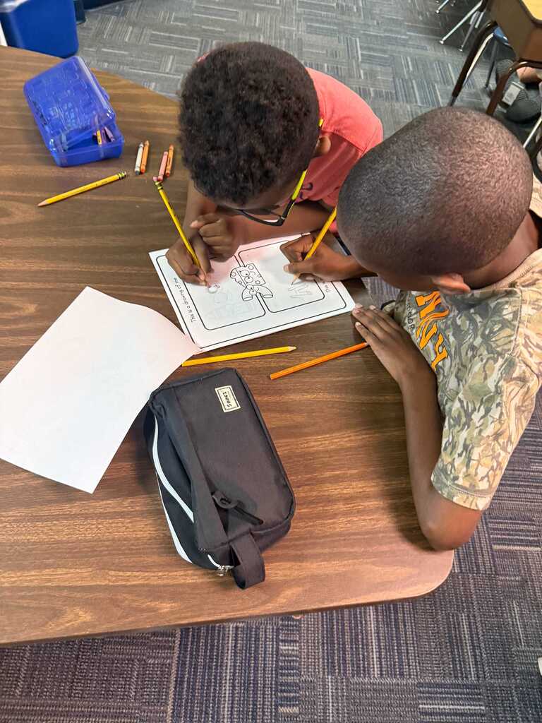 Two children in a classroom setting, each holding pencils, working on a paper with drawings.