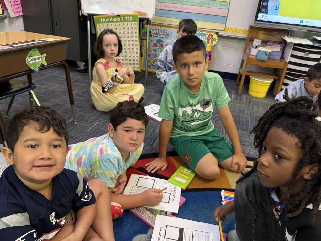 A group of children sitting on a floor in a classroom, with some holding papers. Bright lights and school desks in background.