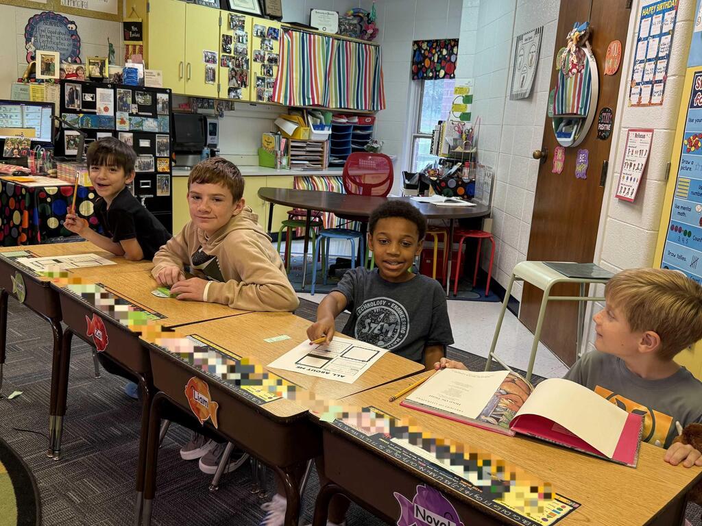 Four children seated at a school desk, engaged in various activities such as reading, writing, and playing with toys.