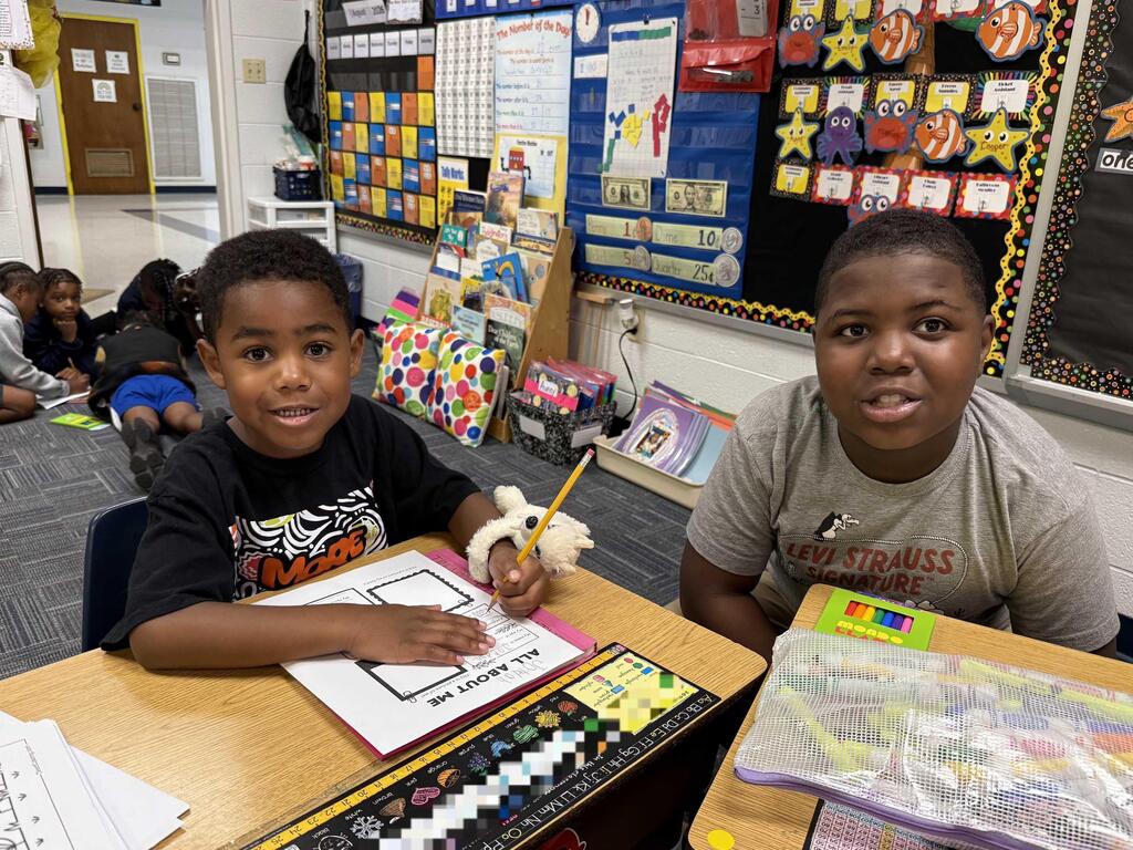 Two children in a classroom. One holds a pencil, focused on a worksheet. Colorful decorations on the wall.