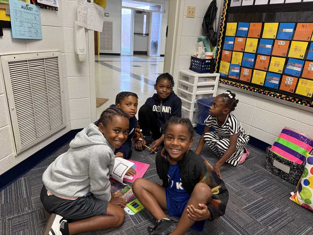 Five children sitting on floor in classroom. One holding book. Brightly colored bulletin board in background.