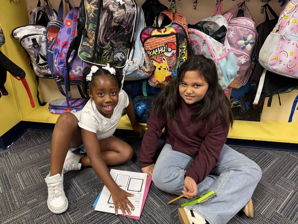 Two children sit on a floor, one holds a book, and the other holds a pencil. They sit in front of colorful backpacks hanging on a wall.