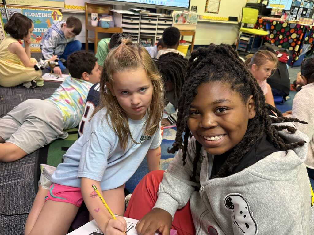 Two girls sit in a classroom, one writing while the other smiles. A diverse group of students are seated around them.