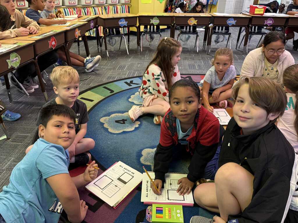 Children sitting on a rug, engaging with educational materials in a classroom. Some are writing, others looking at papers.