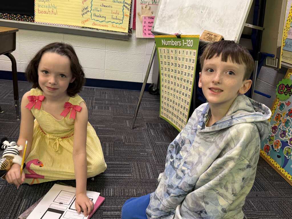 Two children, one in a yellow dress and the other in a hoodie, sit on a floor. Behind them is a whiteboard and a bulletin board with papers.