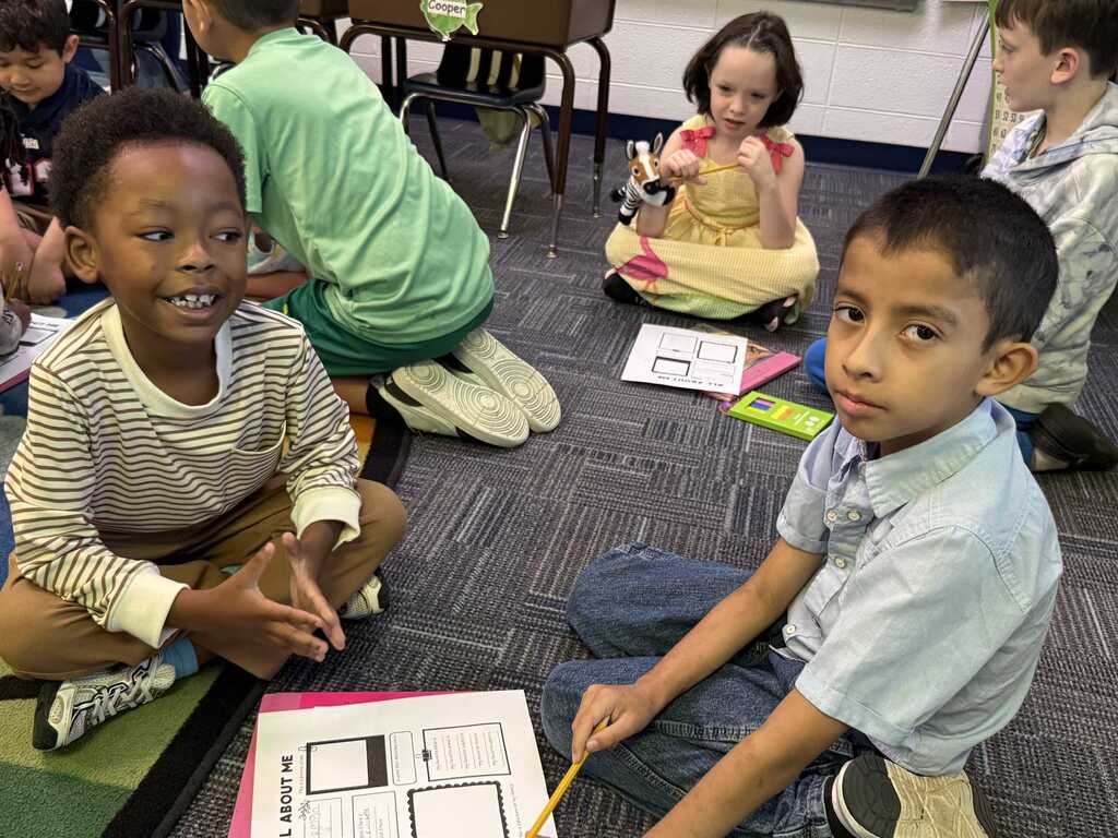 Children seated on floor, engaged in activities; one holds a pencil and looks at a paper.