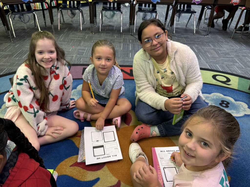 Four children sit on a carpeted floor, engaged in an activity with paper. One holds a paper with shapes.