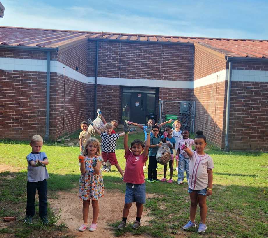 A group of children in a grassy area, with a building in the background. Some children have their hands raised.