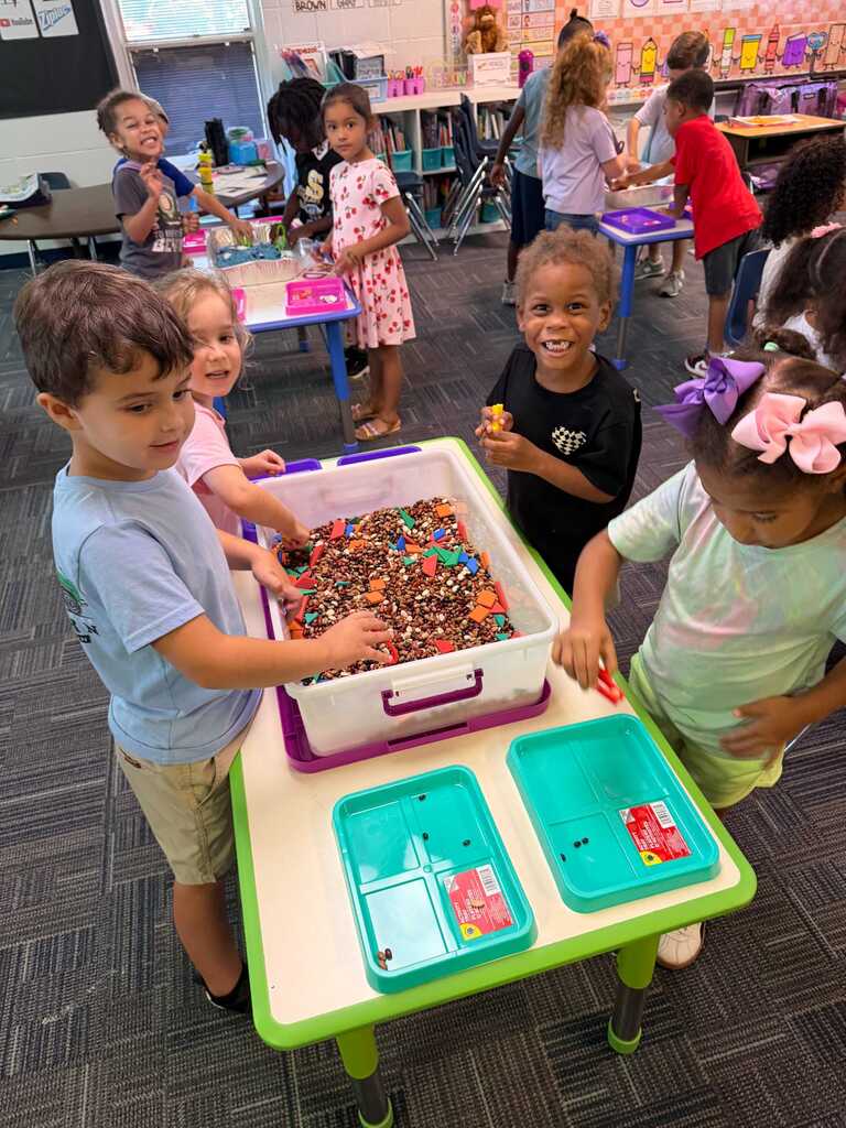Children play with items in a container at a table in a classroom with other children.