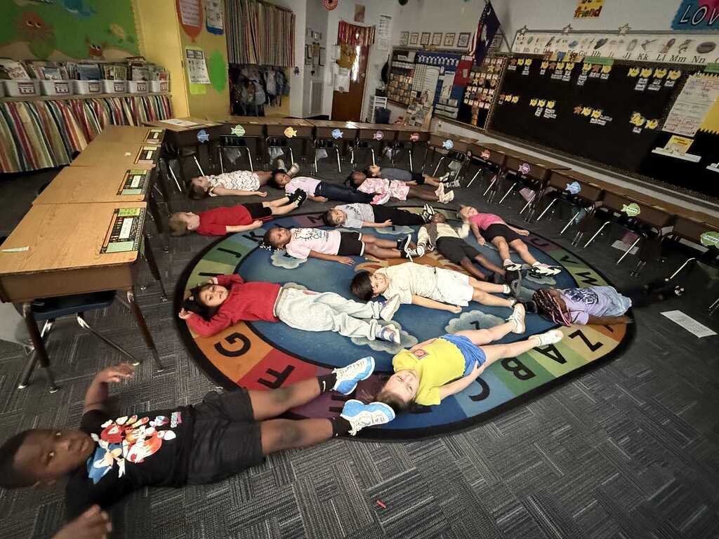 Children lying on a colorful alphabet rug inside a classroom. Desks and chairs are around the room.